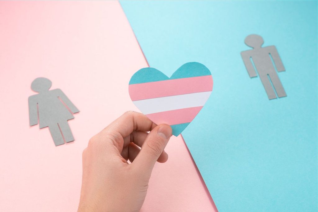 Hand holding a paper heart with transgender pride flag. Paper figures of woman and man on pink and blue background. Trans people and gender identity.